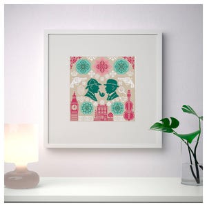 May include: Framed cross-stitch art featuring silhouettes of two men, a pipe, and a violin, with the text "221B". The design includes teal, pink, and beige geometric patterns, guns, and a building. The artwork is displayed in a white frame.