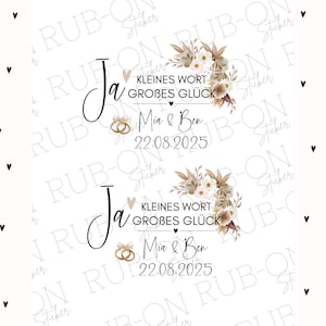 Könnte beinhalten: Hochzeitsaufkleber mit dem Wort "Ja" in Schreibschrift, dem Satz "Kleines Wort, Großes Glück" und den Namen "Mia & Ben" mit dem Datum "22.08.2025." Mit floralen Akzenten und Ehering-Illustrationen.