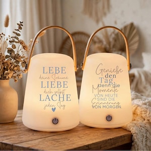 Könnte beinhalten: Zwei weiße, glockenförmige, tragbare LED-Laternen mit Holzgriffen. Die linke Laterne trägt den blauen Text LEBE deine Träume LIEBE dich selbst LACHE jeden Tag. Die rechte Laterne trägt den grauen Text Genieße den Tag, denn die Momente VON HEUTE SIND DIE ERINNERUNG VON MORGEN.