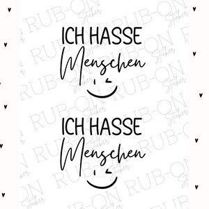 May include: A white sticker featuring the German phrase "ICH HASSE Menschen" in black, translating to "I hate people", with a smiling face. The background is white with small black heart shapes.