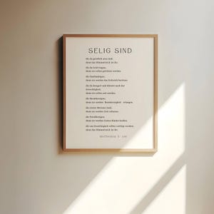 Może przedstawiać: Wydruk w ramie z białym tłem i czarnym tekstem. Tekst to cytat z Biblii, Ewangelii Mateusza 5:3-10, o "Selig Sind" lub "Błogosławieni są" po niemiecku. Cytat jest po niemiecku, a tekst jest wyśrodkowany na stronie.