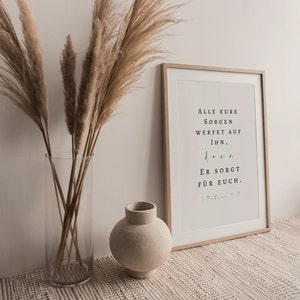 May include: A framed print with a white background and black text. The text reads "ALLE EURE SORGEN WERFET AUF IHN, ER SORGT FÜR EUCH. 1.PETER 5:7". The print is sitting on a beige rug with a vase of dried pampas grass and a small beige ceramic vase.