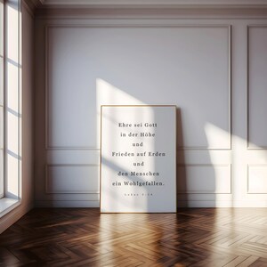 May include: A framed print with gold trim, featuring text in German. The text reads "Ehre sei Gott in der Höhe und Frieden auf Erden und den Menschen ein Wohlgefallen." The print is leaning against a white wall in a room with a wooden floor.