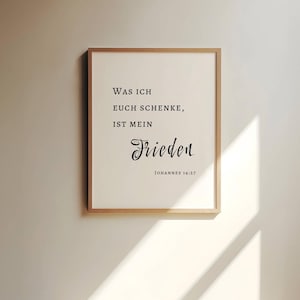 May include: A framed print with the words "Was ich euch schenke, ist mein Frieden" in black script. The print is in a light wood frame and is displayed on a neutral-colored wall. The text is from Johannes 14:27.