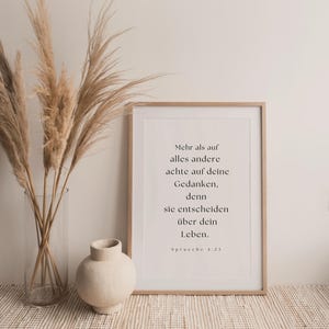 K&ouml;nnte beinhalten: Ein minimalistisches Poster mit wei&szlig;em Hintergrund und schwarzem Text. Der Text lautet: "Mehr als auf alles andere achte auf deine Gedanken, denn sie entscheiden &uuml;ber dein Leben. Spr&uuml;che 4:23". Das Poster ist in einem hellen Holzrahmen gerahmt und steht auf einem Tisch mit einer Vase mit getrocknetem Pampasgras.