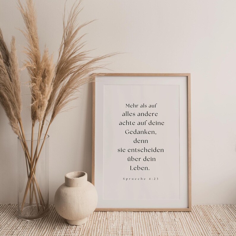 K&ouml;nnte beinhalten: Ein minimalistisches Poster mit wei&szlig;em Hintergrund und schwarzem Text. Der Text lautet: "Mehr als auf alles andere achte auf deine Gedanken, denn sie entscheiden &uuml;ber dein Leben. Spr&uuml;che 4:23". Das Poster ist in einem hellen Holzrahmen gerahmt und steht auf einem Tisch mit einer Vase mit getrocknetem Pampasgras.