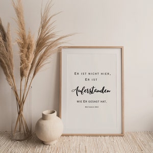May include: A framed print with a white background and black text that reads "ER IST NICHT HIER, ER IST AUFERSTANDEN WIE ER GESAGT HAT. MATTHAEUS 28.6". The print is framed in a light wood frame and is sitting on a light brown woven surface.