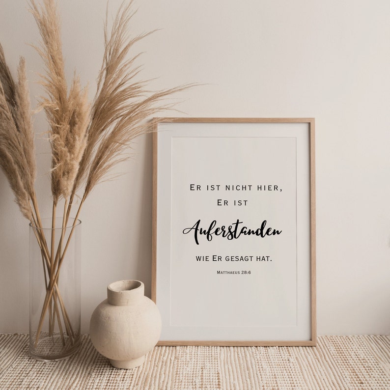 May include: A framed print with a white background and black text that reads "ER IST NICHT HIER, ER IST AUFERSTANDEN WIE ER GESAGT HAT. MATTHAEUS 28.6". The print is framed in a light wood frame and is sitting on a light brown woven surface.