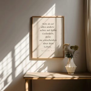 K&ouml;nnte beinhalten: Ein gerahmter Druck mit wei&szlig;em Hintergrund und schwarzem Text. Der Text lautet: "Mehr als auf alles andere achte auf deine Gedanken, denn sie entscheiden &uuml;ber dein Leben. Sprueche 4:23". Der Druck h&auml;ngt an einer Wand mit einer Blumenvase davor.