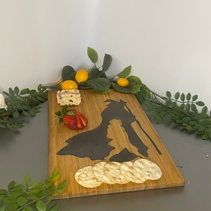 May include: A rectangular bamboo serving board with a dark silhouette design. The board holds crackers, cheese, and a sliced strawberry. Greenery and yellow fruit decorate the edges, creating an appealing presentation for appetizers.