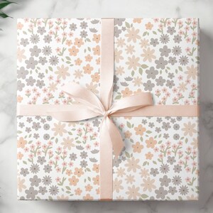 May include: A square gift box wrapped in white paper featuring a repeating pattern of small flowers in shades of peach, light gray, and sage green. The box is tied with a smooth, pale peach satin ribbon arranged in a centered bow.