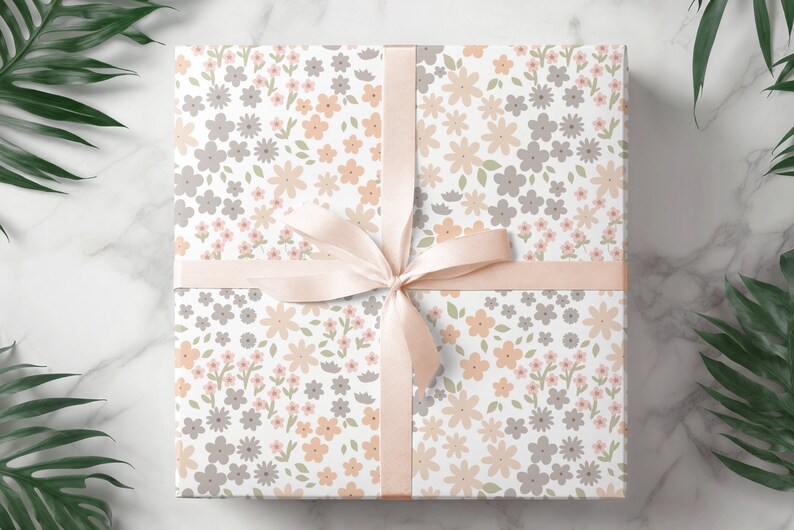 May include: A square gift box wrapped in white paper featuring a repeating pattern of small flowers in shades of peach, light gray, and sage green. The box is tied with a smooth, pale peach satin ribbon arranged in a centered bow.