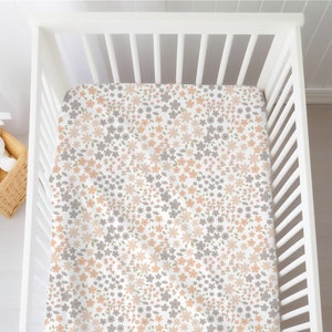 May include: A white crib sheet featuring a repeating floral pattern in shades of muted peach, soft grey, and light green. The fabric is fitted over a white slatted crib mattress base, creating a clean and consistent look for the bedding surface.