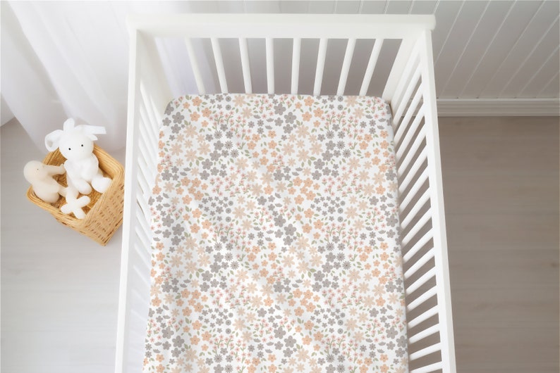 May include: A white crib sheet featuring a repeating floral pattern in shades of muted peach, soft grey, and light green. The fabric is fitted over a white slatted crib mattress base, creating a clean and consistent look for the bedding surface.