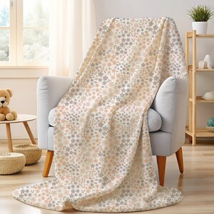 May include: A soft throw blanket featuring a repeating pattern of small, delicate flowers in muted shades of peach, light gray, and white. The fabric has a plush texture and is draped over a light gray armchair, covering the seat and backrest.