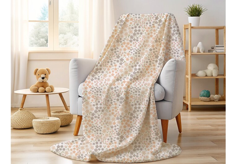 May include: A soft throw blanket featuring a repeating pattern of small, delicate flowers in muted shades of peach, light gray, and white. The fabric has a plush texture and is draped over a light gray armchair, covering the seat and backrest.