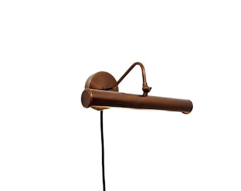 May include: A wall-mounted picture light with a cylindrical bronze-toned metal housing. The fixture features an adjustable arm connected to a circular wall plate and includes a thin black power cord extending downward from the base.