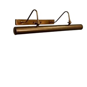 May include: A brass picture light, featuring a rectangular backplate and a cylindrical light housing. Two curved arms connect the backplate to the light. This vintage-style light fixture is designed to illuminate artwork, with a warm, inviting glow.