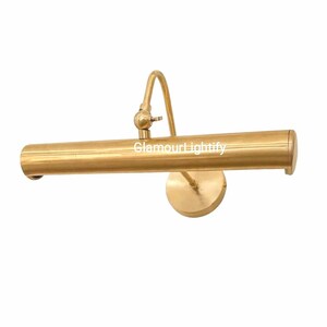 May include: A brass picture light with a cylindrical lamp head and a curved arm. The light is mounted on a round base and has a vintage aesthetic. The text "GlamourLightify" is visible.