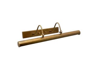 May include: A brass picture light fixture featuring a long, cylindrical horizontal tube attached to a rectangular wall-mounting plate by two curved, adjustable metal arms. The metal has a smooth, matte finish with a warm, golden-brown tone.