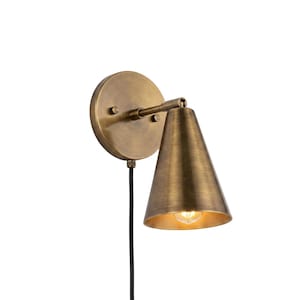 May include: A wall-mounted lamp featuring a circular brass backplate and a conical brass shade. The fixture includes a short horizontal arm connecting the base to the shade and a black power cord extending downward from the bottom of the backplate.