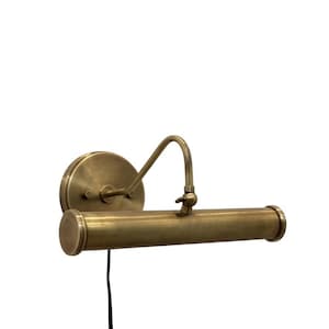 Puede incluir: Una lámpara de pared para cuadros con acabado en latón antiguo. El aplique cuenta con una placa de montaje circular, un brazo curvo y una carcasa de luz cilíndrica horizontal. Un cable de alimentación negro y fino cuelga hacia abajo.