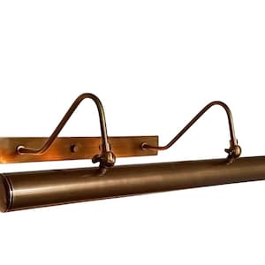 May include: A bronze picture light. The light has a rectangular backplate, curved arms, and a cylindrical light housing. The fixture is designed to illuminate artwork. The light is mounted on a white background.