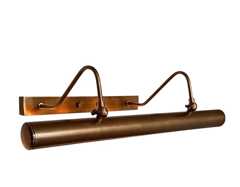 May include: A bronze picture light. The light has a rectangular backplate, curved arms, and a cylindrical light housing. The fixture is designed to illuminate artwork. The light is mounted on a white background.