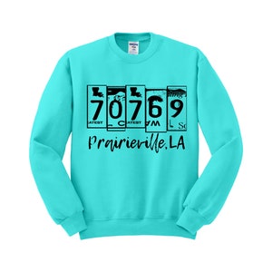 May include: Turquoise blue sweatshirt with a black graphic print of a vintage-style license plate with the numbers 70769 and the text "Prairieville, LA".