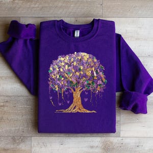 May include: Purple sweatshirt with a gold tree design. The tree is made up of small gold, purple, and green leaves. The tree has a gold trunk and branches. The tree is surrounded by small gold hearts.
