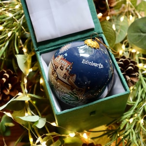 May include: A blue glass Christmas ornament with a painted scene of Edinburgh, Scotland. The ornament is in a green velvet box with a white satin lining. The ornament has gold accents and the text "Edinburgh" is written on the front.