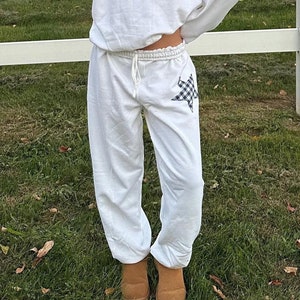 May include: White sweatpants with a black and white checkered star on the left leg. The sweatpants are paired with a white sweatshirt with a black and white checkered star on the front. The model is wearing brown suede boots.