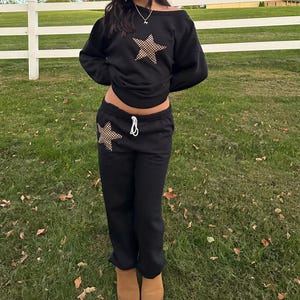 May include: Black sweatshirt with a star design and matching black sweatpants with a star design on the leg. The outfit is worn by a person.