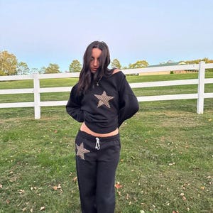 May include: A black sweatshirt with a star design and black sweatpants with a star design. The model is wearing the outfit and standing in front of a white fence.
