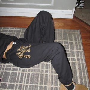 May include: Black sweatpants with a leopard print New York Yankees logo on the left leg. The sweatpants are on a gray and white checkered rug.