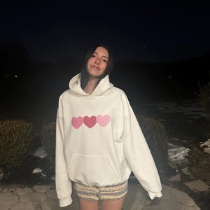 May include: A white hoodie with three pink hearts on the front. The hearts are made of a patchwork fabric with pink and white squares.