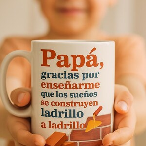 Puede incluir: Taza de cerámica blanca con el texto en español "Papá, gracias por enseñarme que los sueños se construyen ladrillo a ladrillo" en rojo y azul. La taza presenta una ilustración de pared de ladrillos y llana.