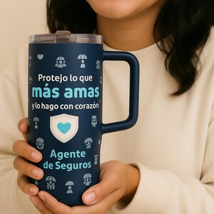 以下が含まれることがあります： ハンドル付きの濃いネイビーブルーのトラベルマグ。テキスト「Protejo lo que más amas y lo hago con corazón」と「Agente de Seguros」が特徴です。マグには蓋があり、小さな傘とハートのアイコンで飾られています。