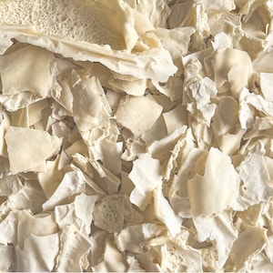 Op de afbeelding: Een close-up van een stapel gedroogde, witte en gebroken witte, onregelmatig gevormde, papierachtige stukken. De stukken zijn verspreid en overlappen elkaar.