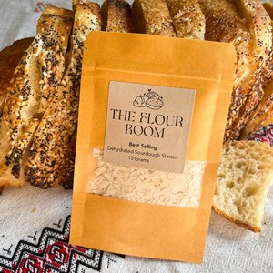 May include: A small brown paper bag with a white label that says "THE FLOUR ROOM" and "Best Selling Dehydrated Sourdough Starter 15 Grams". The bag is sitting on a white and red patterned tablecloth next to a loaf of sliced bread.
