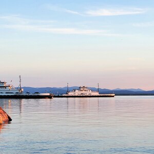May include: Two white ferries moored at a rocky shore with a mountain range in the background. The sky is a soft pink and blue.