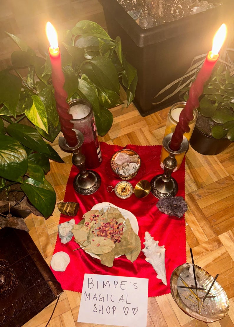K&ouml;nnte beinhalten: Ein roter Samtaltartuch mit zwei brennenden roten Kerzen, einem Kompass, Kristallen und einer Schale mit Lorbeerbl&auml;ttern und Rosenbl&uuml;ten. Ein handgeschriebener Zettel lautet "Bimpe's Magical Shop ♡♡".