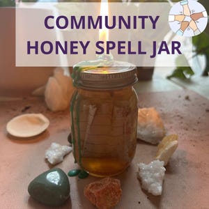 Op de afbeelding: Een glazen pot gevuld met honing en afgesloten met een metalen deksel. De pot is omringd door verschillende kristallen en stenen, waaronder een groene steen, een rode steen en verschillende witte stenen. De pot wordt verlicht door een kaarsvlam, en de tekst "COMMUNITY HONEY SPELL JAR" is zichtbaar boven de pot.