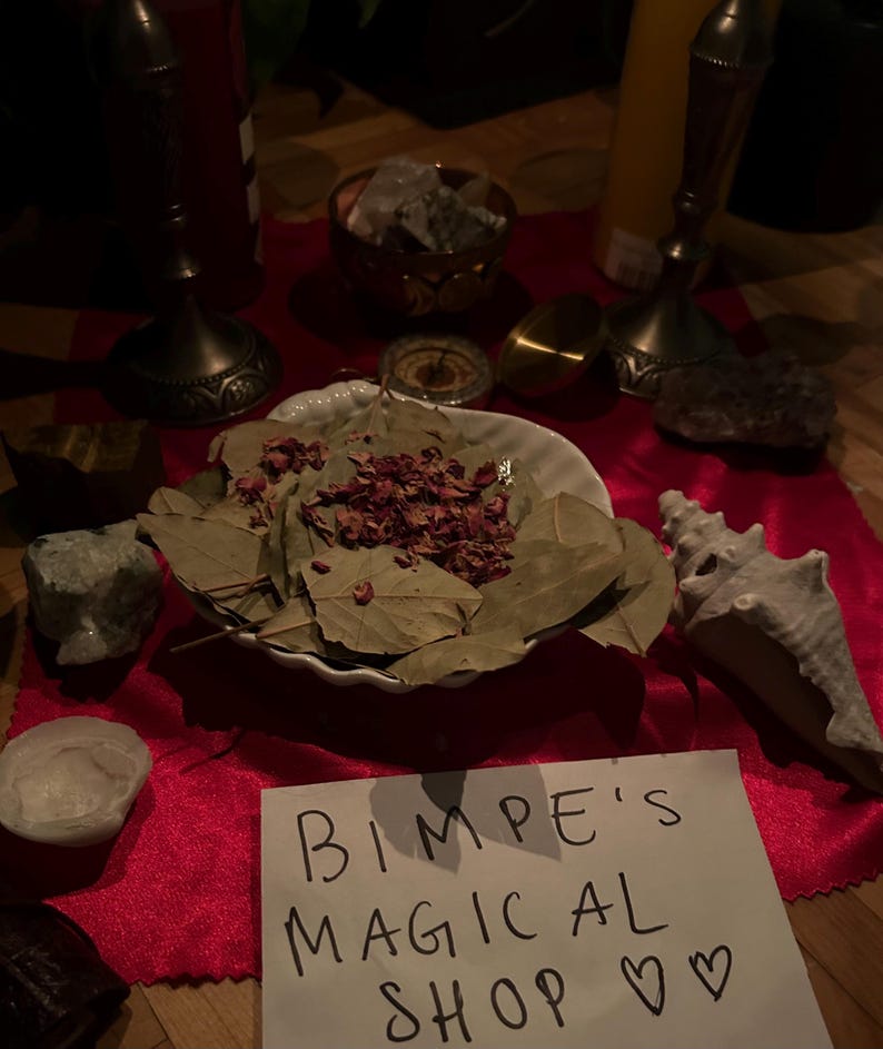 May include: A white bowl filled with dried bay leaves and rose petals sits on a red velvet surface. A seashell and a small white stone are also on the surface. A handwritten sign reads "Bimpe's Magical Shop ♡".