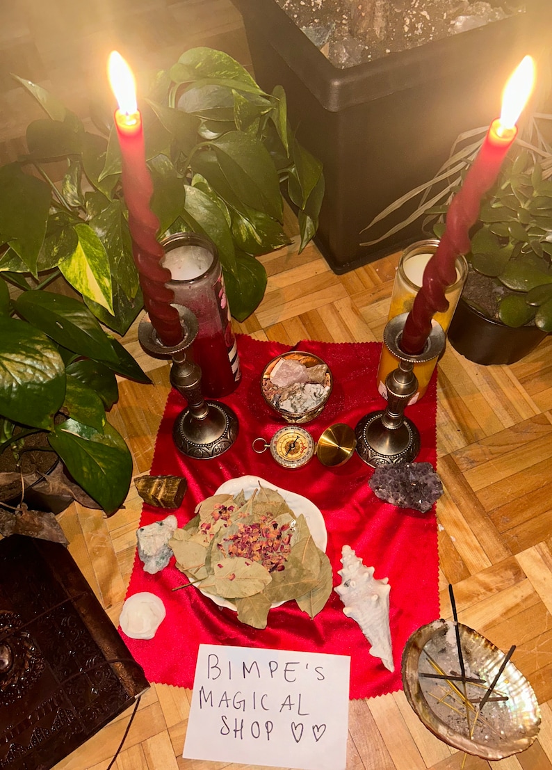 Puede incluir: Un pa&ntilde;o de altar de terciopelo rojo con dos velas encendidas, una br&uacute;jula, cristales, una concha, hojas de laurel y una nota escrita a mano que dice "Bimpe's Magical Shop &hearts;&hearts;".