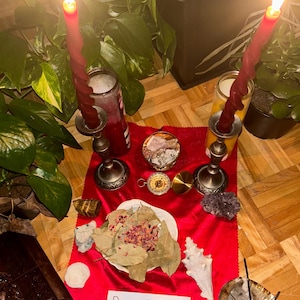May include: A spiritual display with two burning red candles in decorative holders, set on a red cloth. The scene includes crystals, a compass, a seashell, and a handwritten sign reading "BIMPE'S MAGICAL SHOP ♡♡".