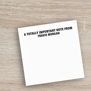 以下が含まれることがあります： 白いキーボード、コーヒーカップ、そして「A TOTALLY IMPORTANT NOTE FROM TRAVIS MORGAN」というテキストのメモ帳が、明るい木目の表面にあります。白いイヤホンが見えます。
