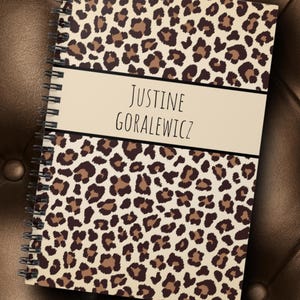 Puede incluir: Un cuaderno de espiral con una cubierta de estampado de leopardo. La cubierta tiene un fondo crema con manchas marrones y negras. El cuaderno tiene una etiqueta de color crema con el nombre "JUSTINE GORALEWICZ" impreso en negro.