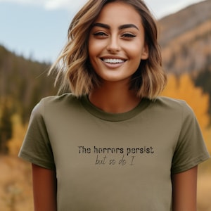 May include: A woman wearing a green t-shirt with the text "The horrors persist but so do I" printed on it.