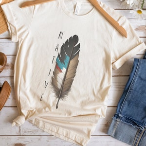 May include: Cream-colored t-shirt with the word "NATIVE" printed vertically on the left side. A detailed feather design in shades of gray, brown, blue, and red is centered on the shirt. The shirt is hanging on a wooden hanger.